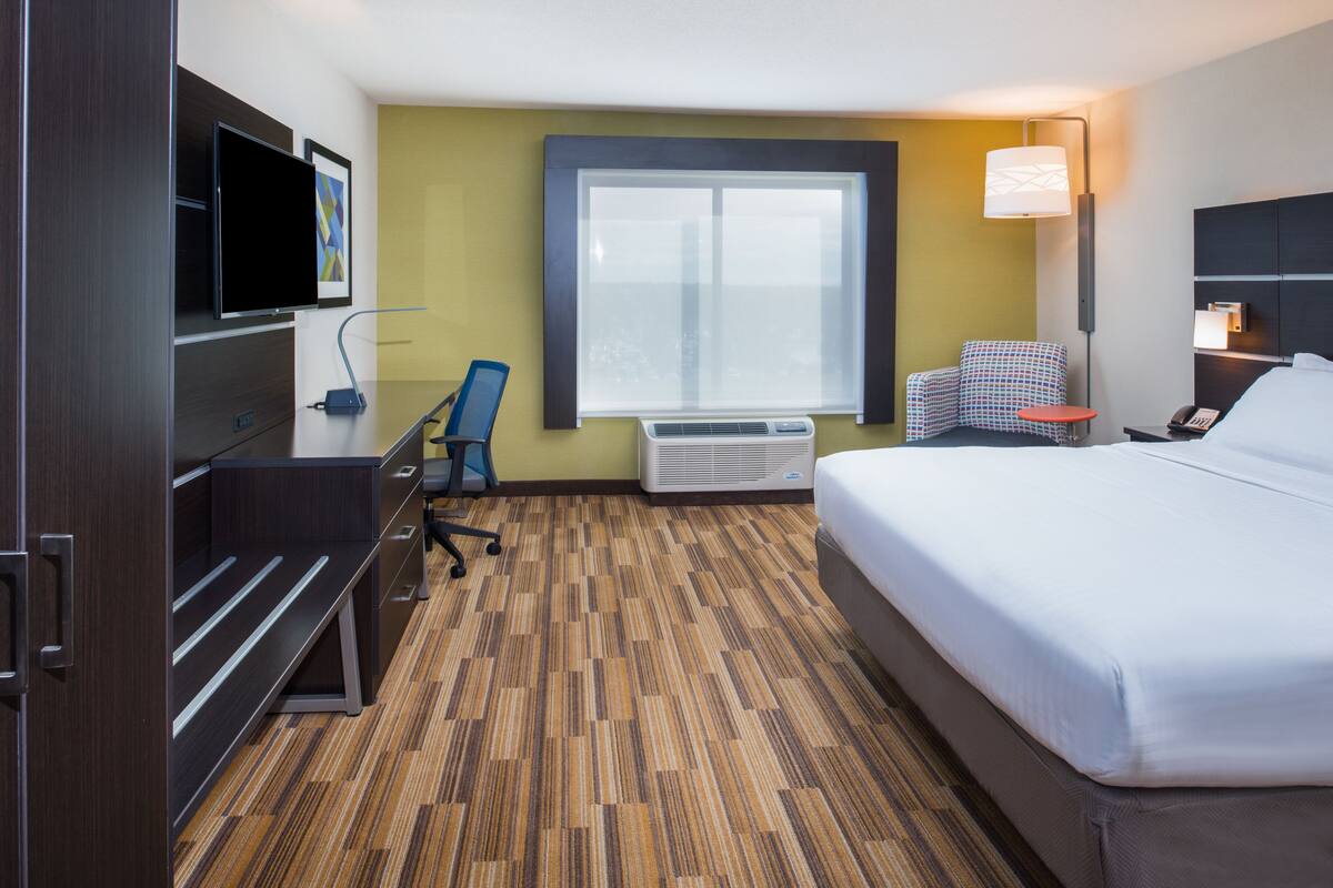 A hotel room with a bed, desk, chair, TV, air conditioning unit, and a window.
