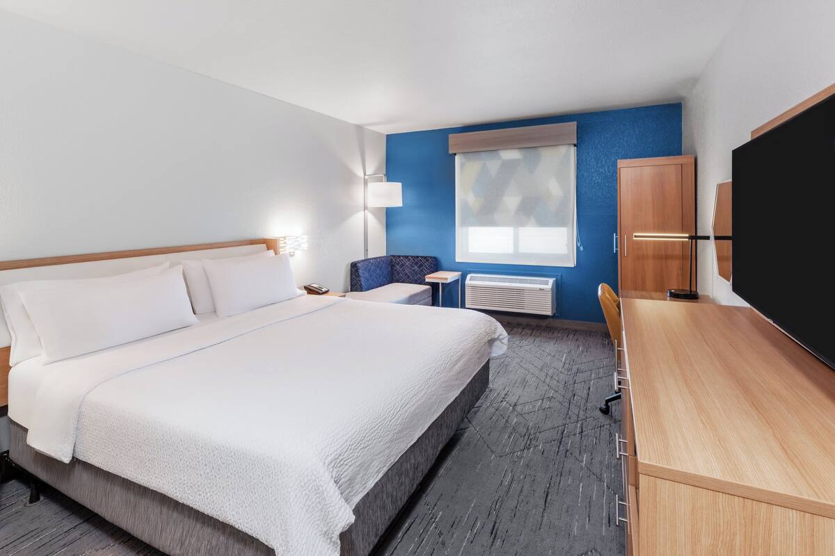 A hotel room with a large bed, a desk, a chair, a window with blinds, a lamp, and a wardrobe.