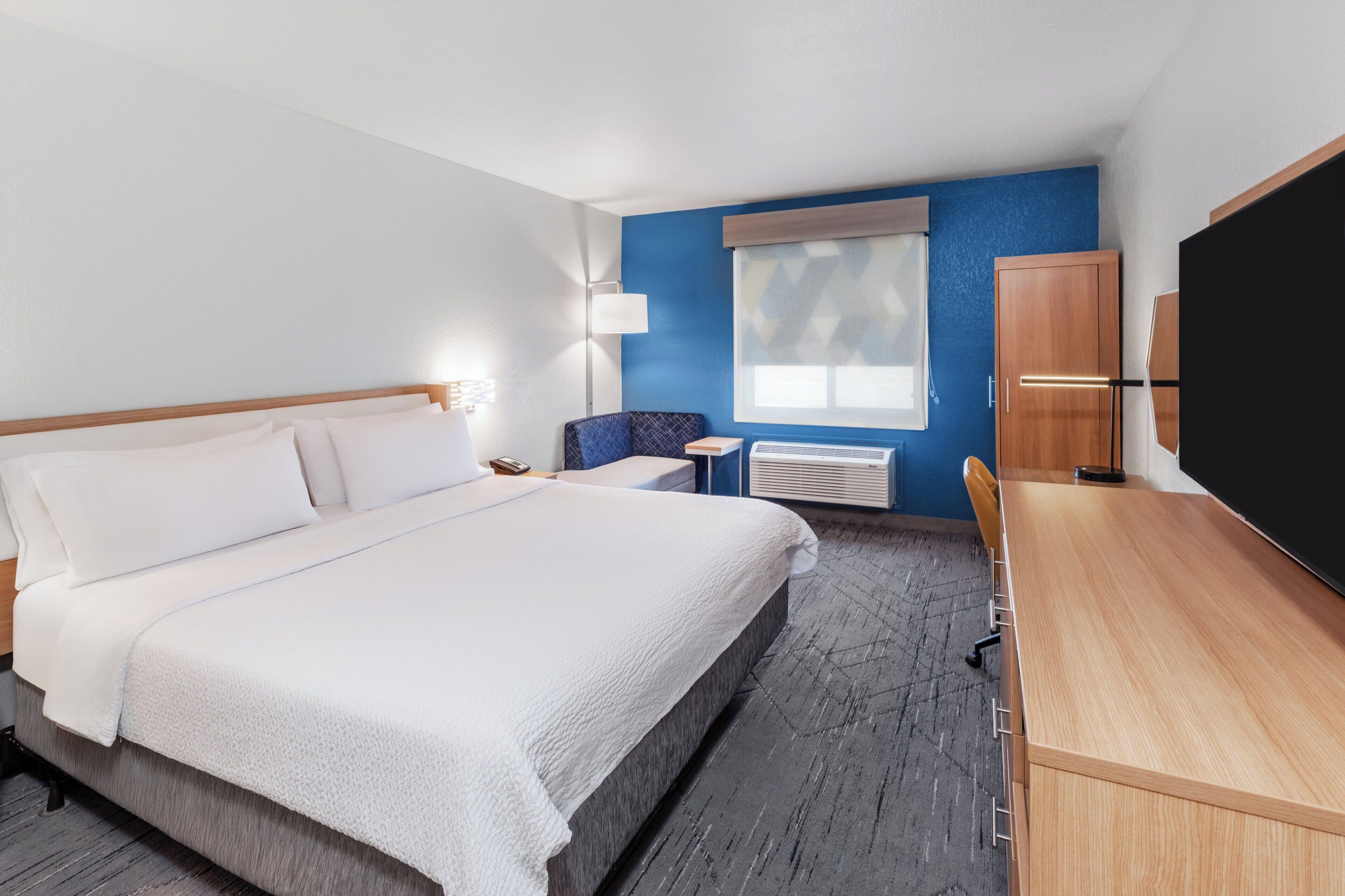 A hotel room with a large bed, a desk, a chair, a window with blinds, a lamp, and a wardrobe.