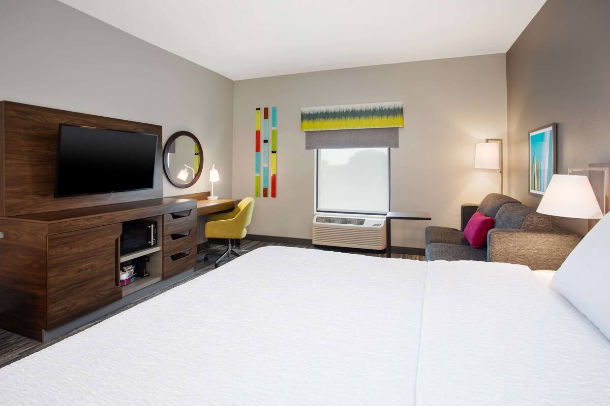 A hotel room with a large bed, a flat-screen TV, a desk with a chair, a window with a view, and a wall-mounted air conditioning unit.