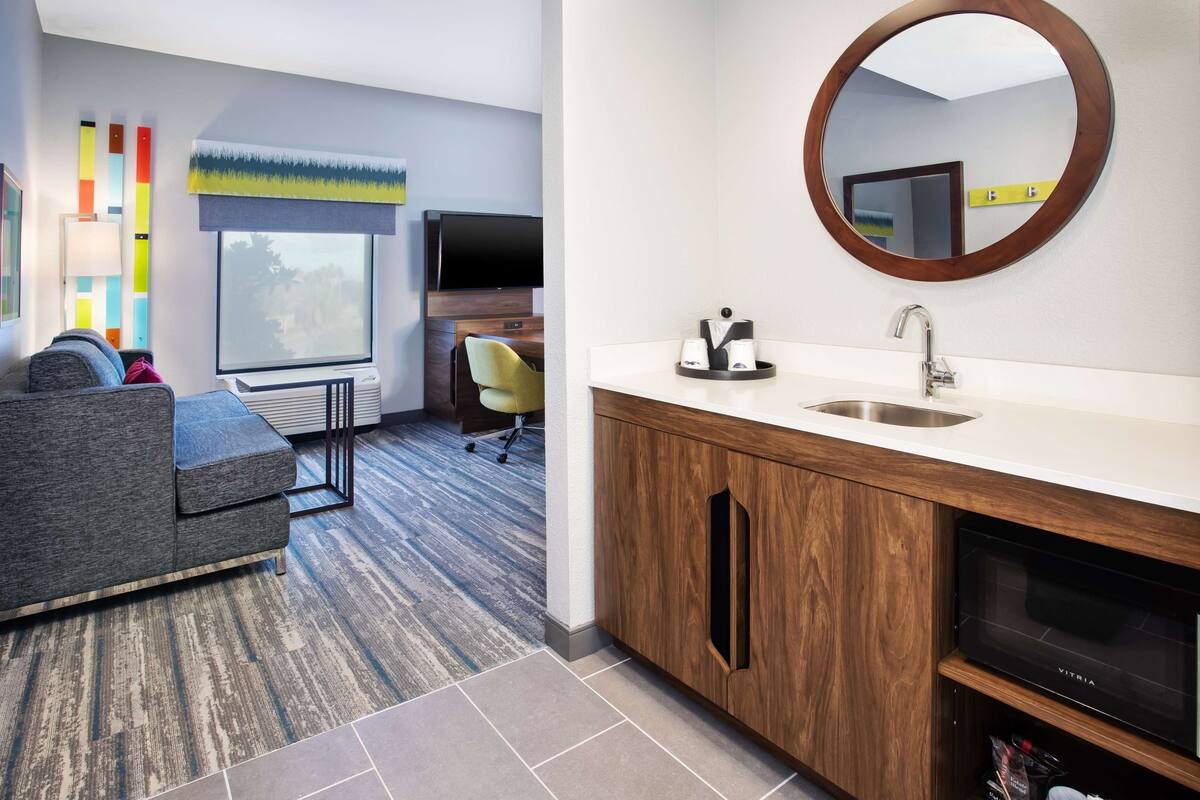 A modern hotel room with a sofa, a TV, a wooden cabinet with a sink, and a large window.