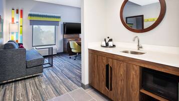 A modern hotel room with a sofa, a TV, a wooden cabinet with a sink, and a large window.