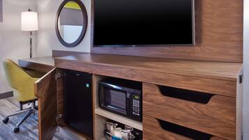 A hotel room with a wooden desk, a flat-screen TV, a microwave, a mini-fridge, a yellow chair, and a lamp.