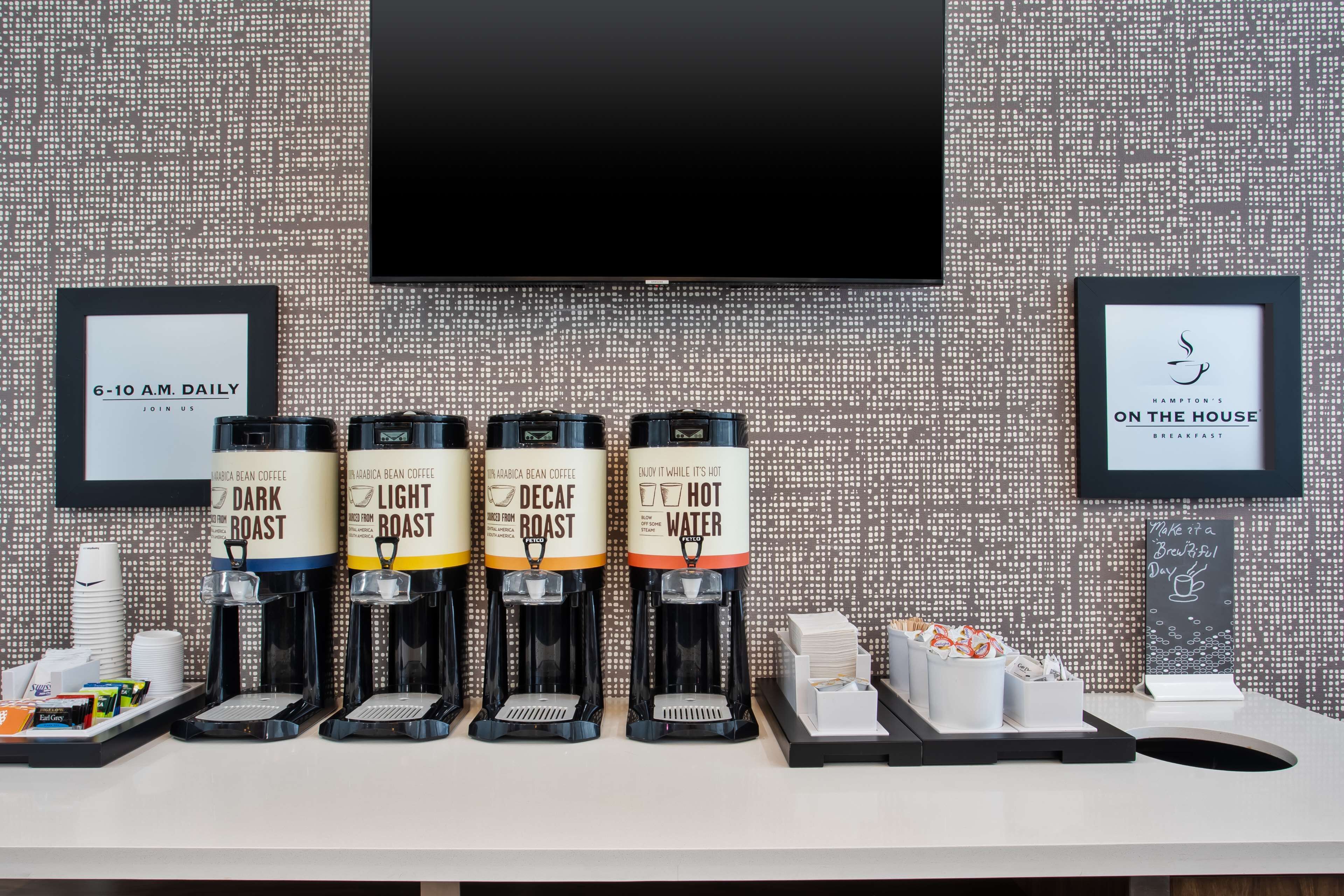 Un espace dédié au café, équipé de quatre distributeurs de café étiquetés « Mélange corsé », « Mélange léger », « Décaféiné » et « Eau chaude ». On y trouve également des tasses à café et un plateau avec des sachets de sucre.