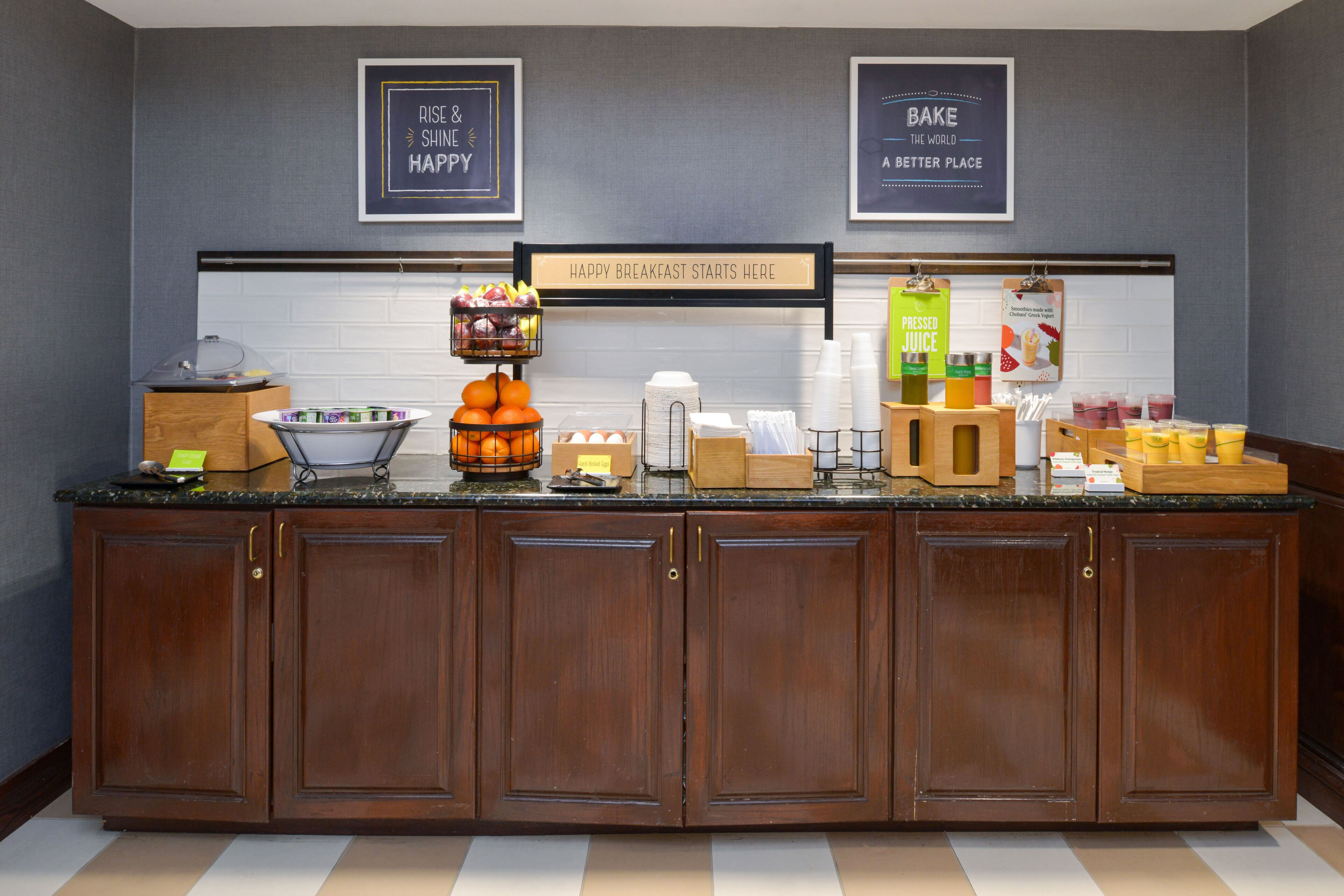 Una barra de desayuno con un cartel que dice “Happy Breakfast Starts Here”. Se exhiben diferentes alimentos y bebidas, entre ellos frutas, jugos y pasteles.