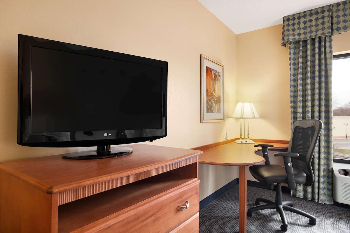 Quarto de hotel com televisão de ecrã plano sobre uma secretária de madeira, uma cadeira, um candeeiro e uma janela com cortinas.