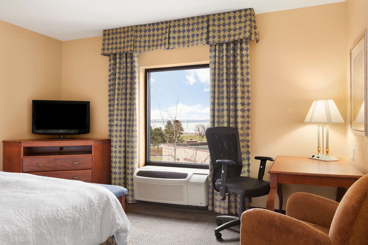 Quarto de hotel com cama, escrivaninha com luminária, cadeira, televisão e janela com cortinas.