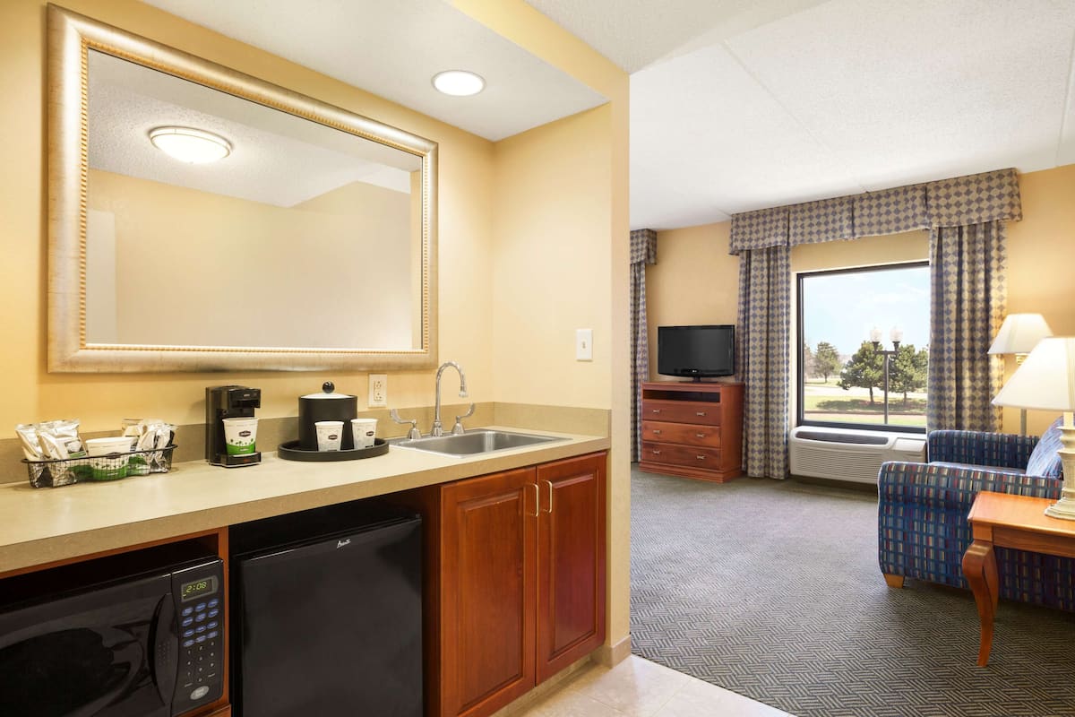 Quarto de hotel com cozinha compacta, um espelho grande, televisão, sofá e janela com cortinas.