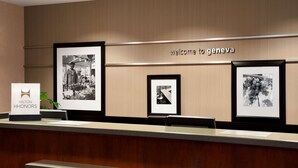 Un comptoir d’accueil d’hôtel avec des photographies encadrées accrochées au mur, une pancarte indiquant « Bienvenue à Genève » et un panneau « Hilton Honors ».