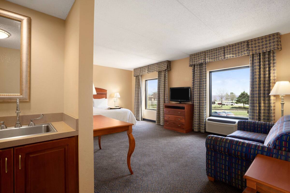 Quarto de hotel com cama, escrivaninha, cadeira, televisão e janela com cortinas.