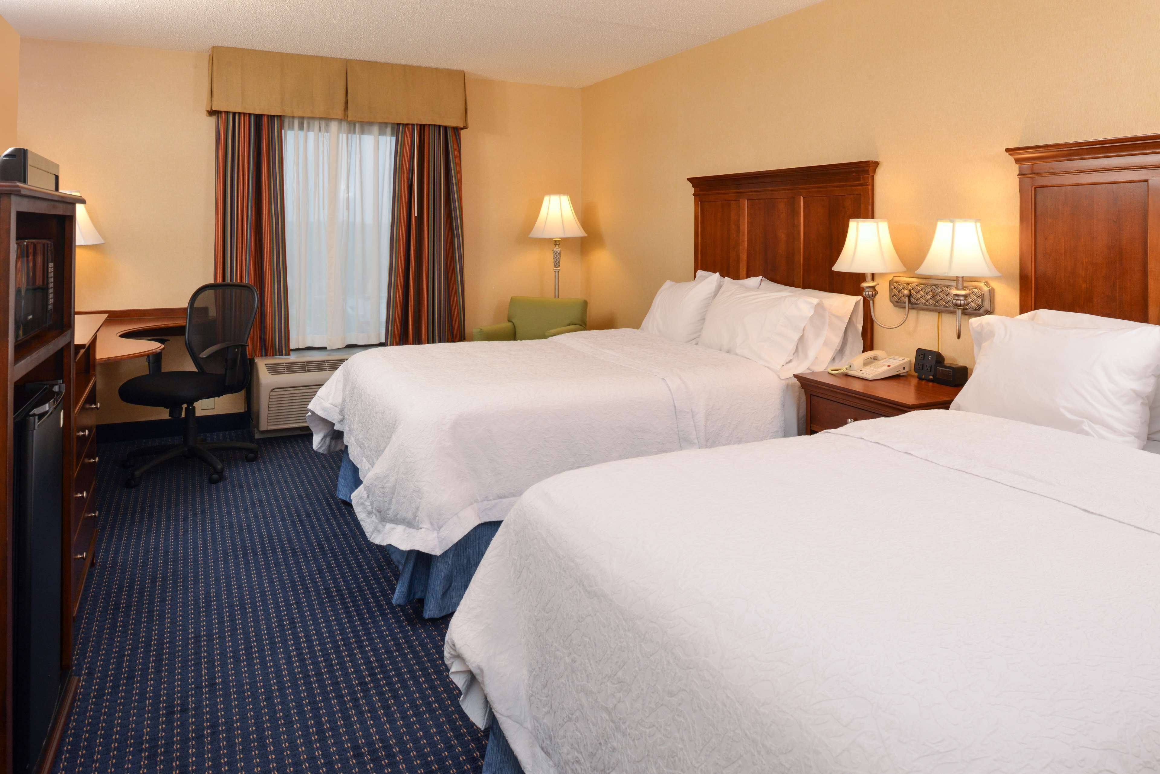 Ein Hotelzimmer mit zwei Betten, einem Schreibtisch mit Computer, einem Sessel und einem Fenster mit Vorhängen.