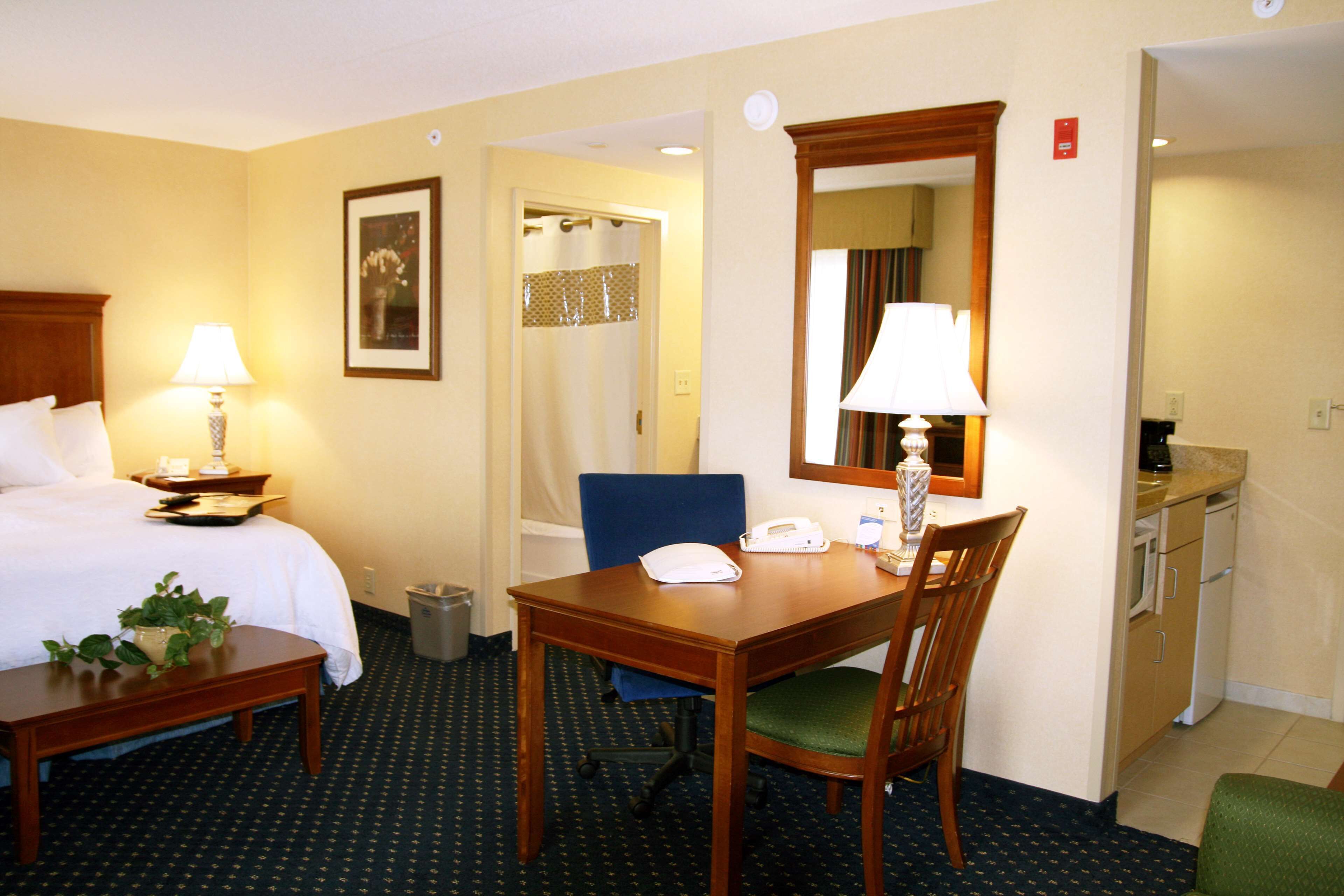 Habitación de hotel con cama, mesita de noche, lámpara, silla, escritorio y una pequeña zona de kitchenette.