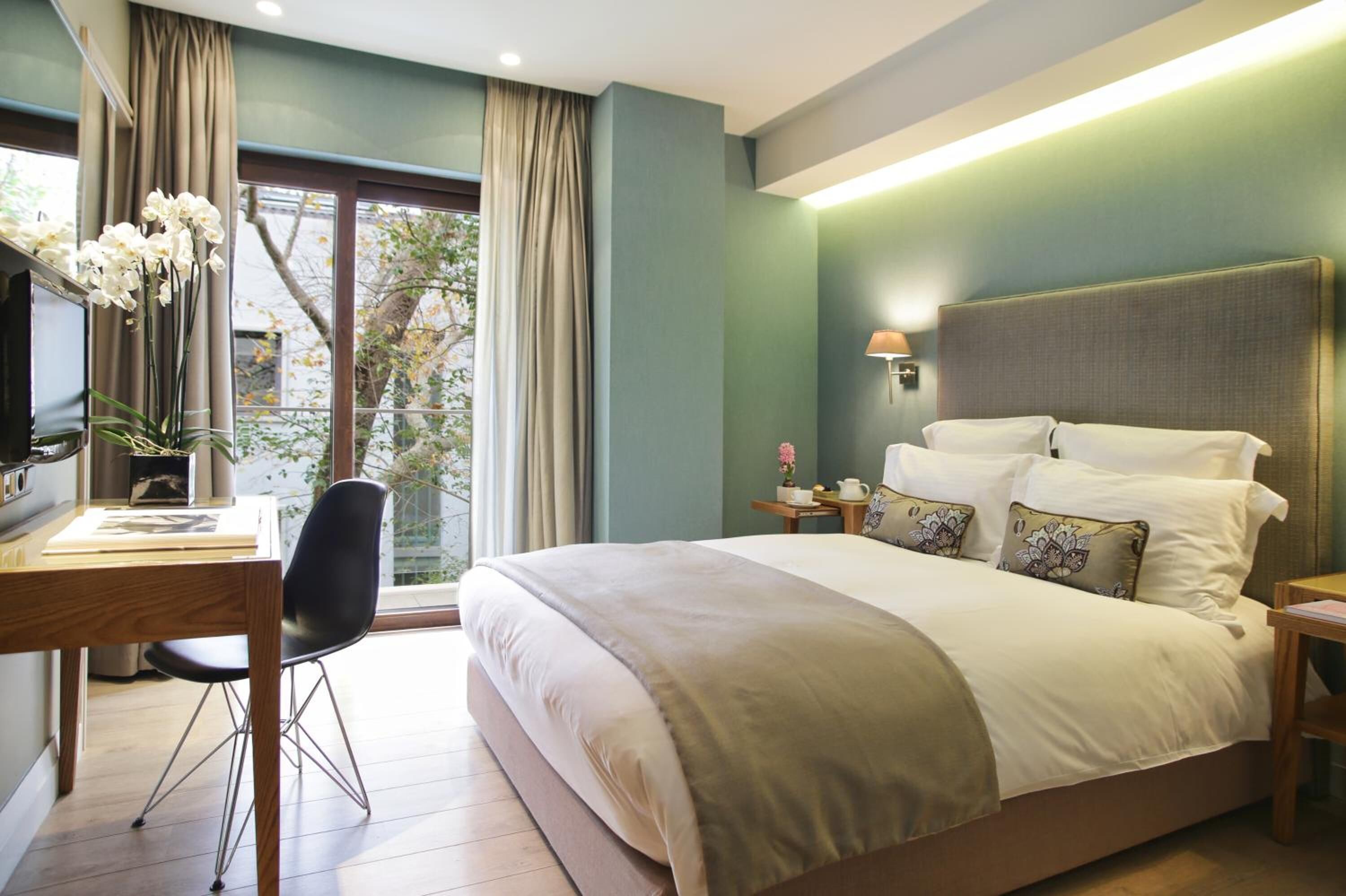 A modern hotel room with a large bed, a desk with a chair, a lamp, and a view of the outdoors.