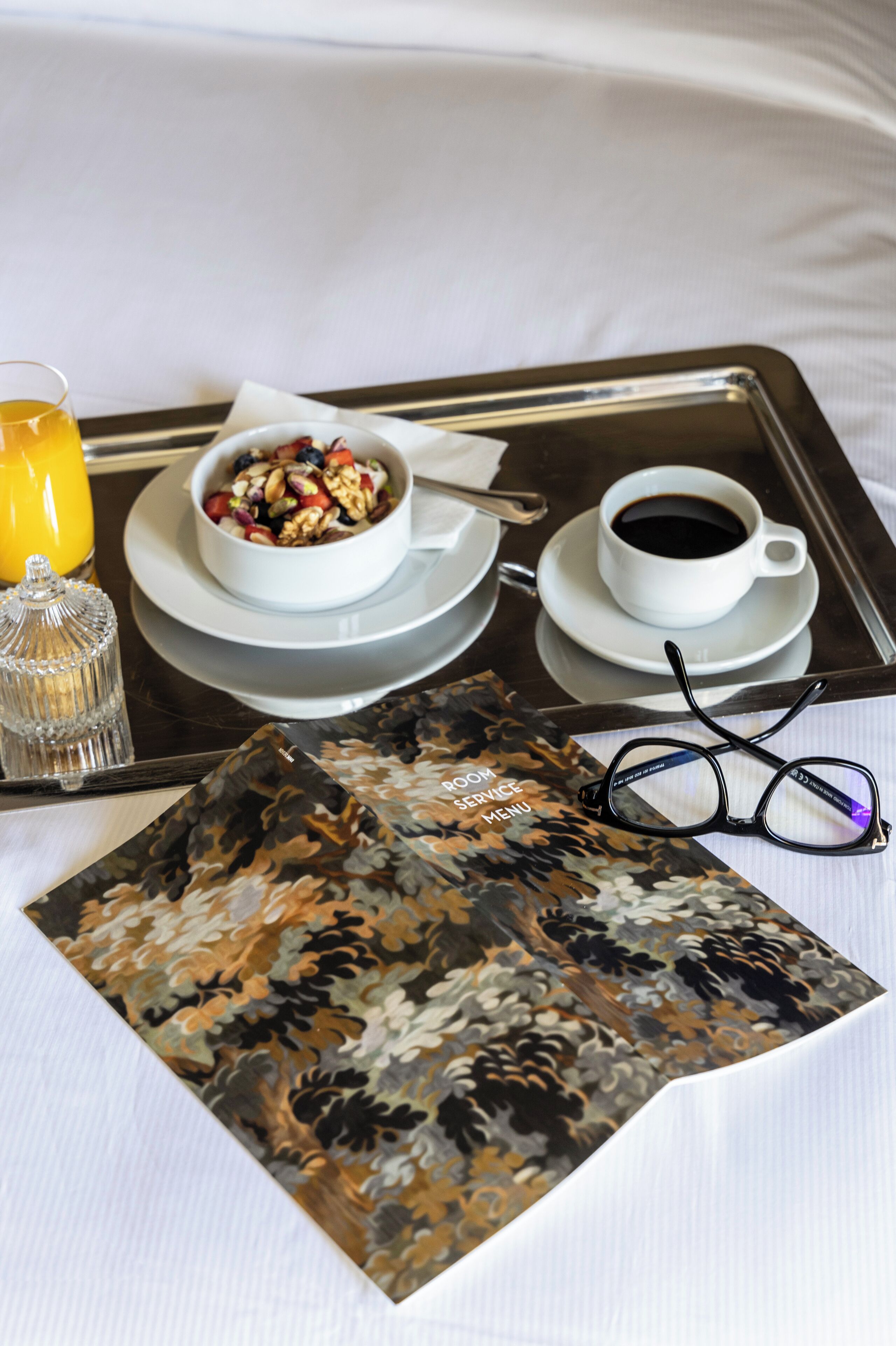 A tray with a bowl of cereal, a cup of coffee, a glass of orange juice, a pair of glasses, and a menu on a bed.
