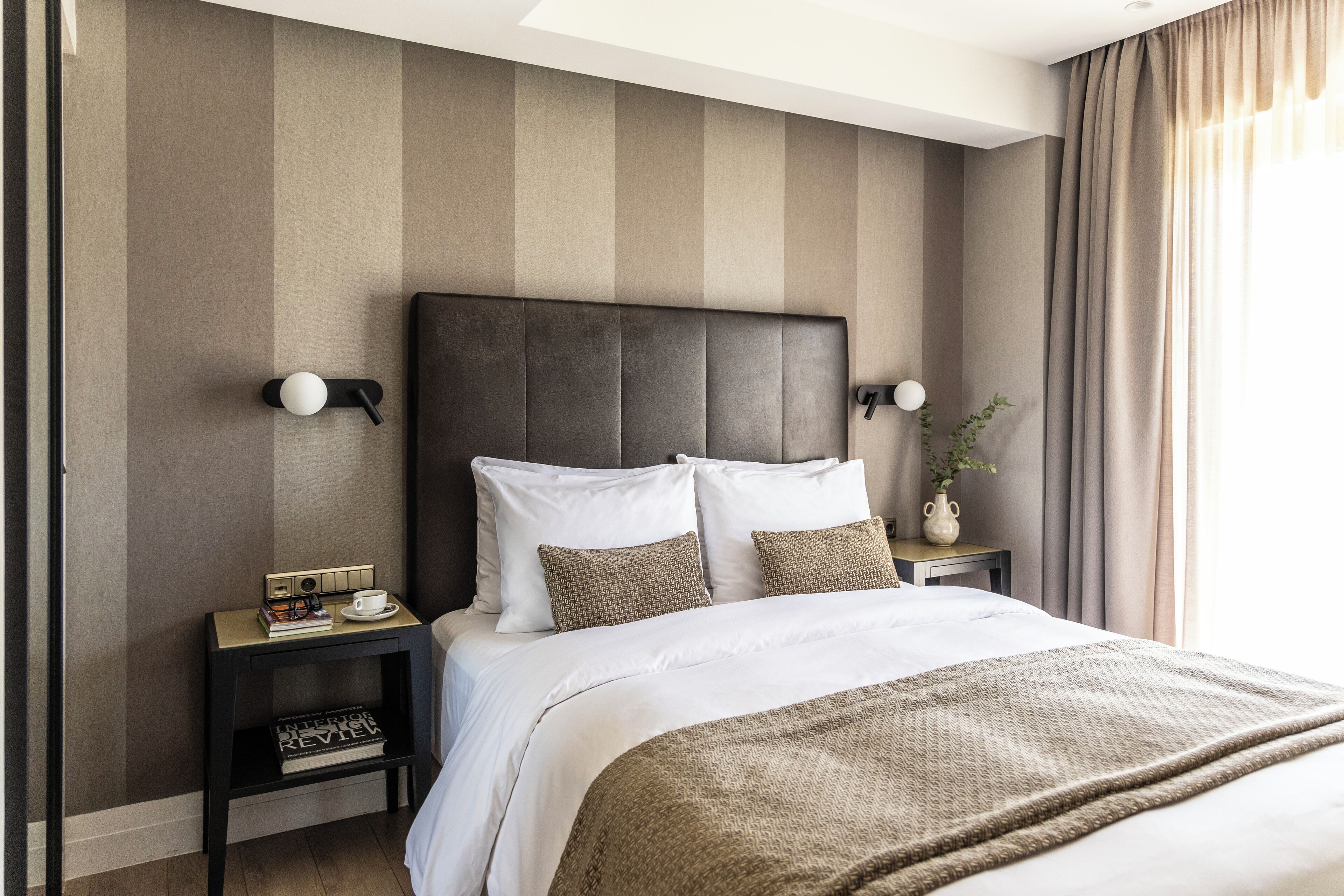 Quarto de hotel com uma cama grande, duas mesas de cabeceira, um criado-mudo, uma luminária na parede e uma janela com cortinas.