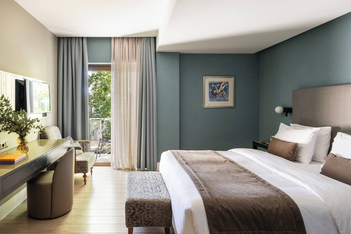 A hotel room with a bed, a desk, a chair, a bench, a TV, and a window with curtains.