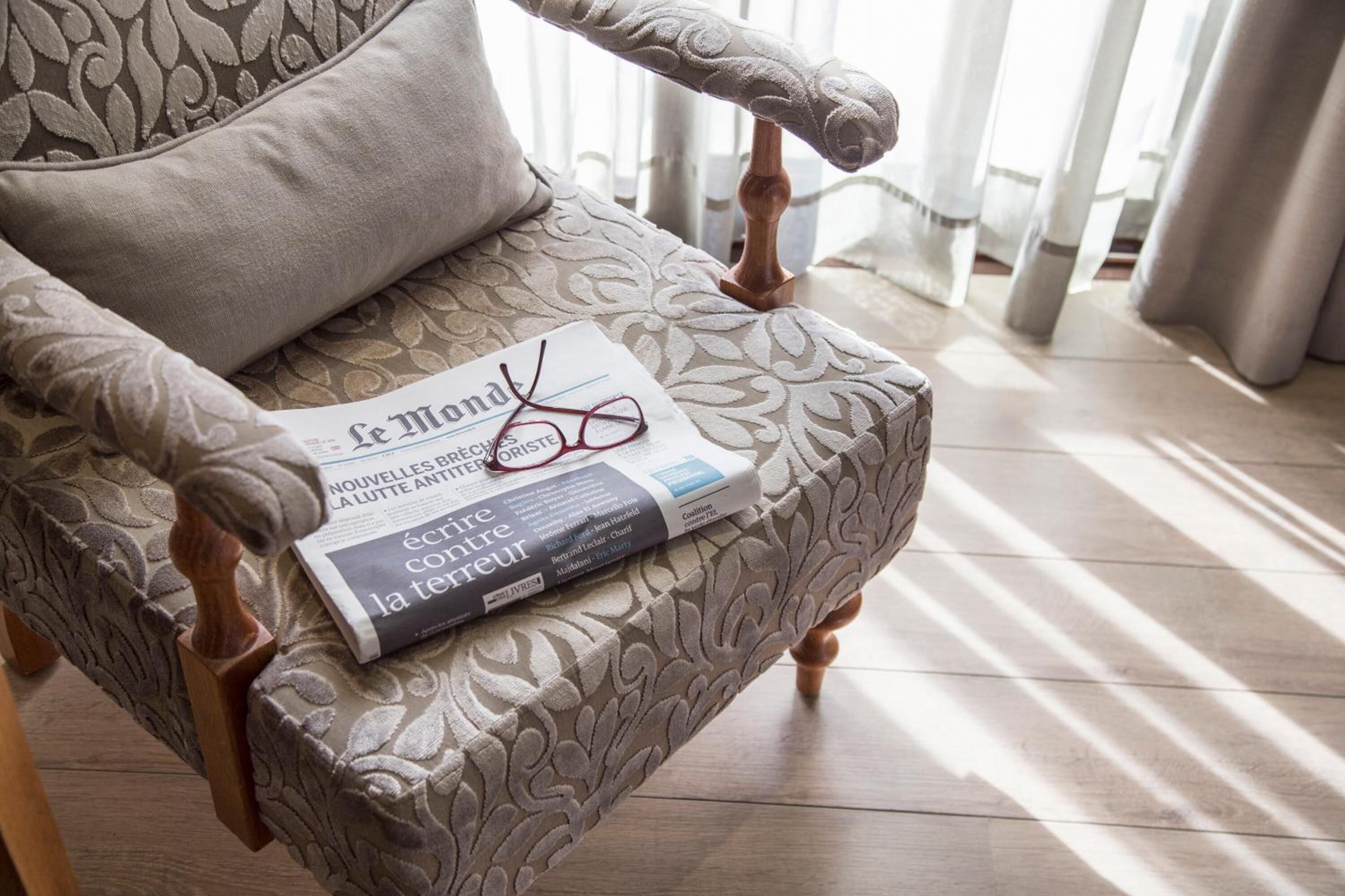 Uma cadeira com tecido estampado e apoio de braço de madeira, um travesseiro, uma revista e um par de óculos.