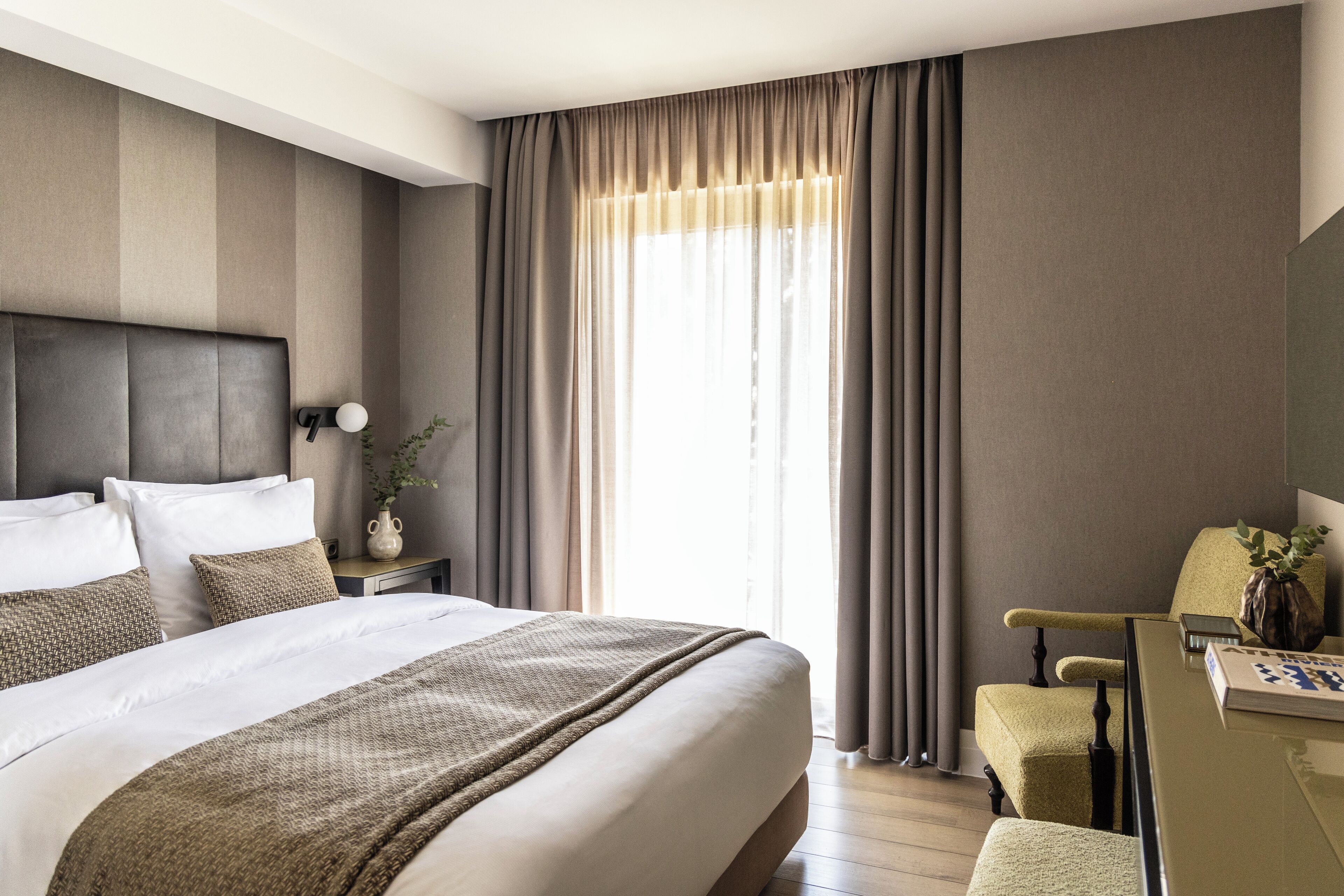 Une chambre d’hôtel avec un grand lit, un bureau et une chaise. Il y a une fenêtre avec des rideaux, une table de chevet avec une lampe et un vase avec une plante.