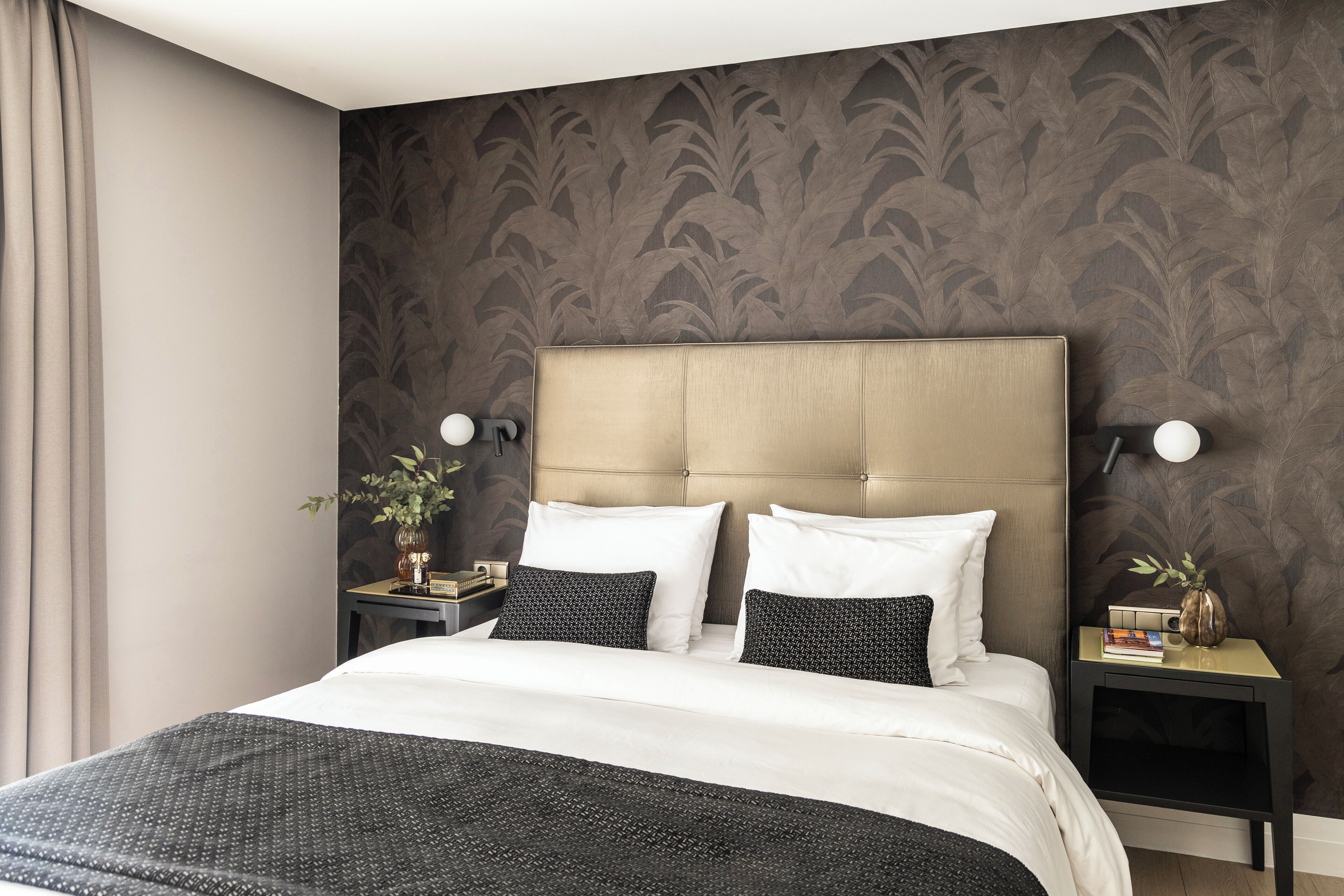 A neatly made bed with a white and black patterned bedspread, two white pillows, and a black throw blanket. There are two bedside tables with lamps, a vase with a plant, and a small stack of books. The room has dark patterned wallpaper and a beige headboard.