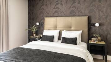 Una cama bien hecha con una colcha de diseño blanco y negro, dos almohadas blancas y un plaid negro. Hay dos mesitas de noche con lámparas, un florero con una planta y una pequeña pila de libros. La habitación tiene papel tapiz de diseño oscuro y un cabecero color beige.