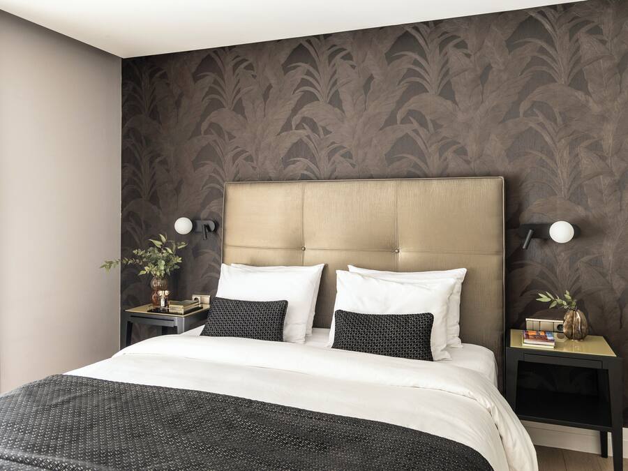 Una cama bien hecha con una colcha de diseño blanco y negro, dos almohadas blancas y un plaid negro. Hay dos mesitas de noche con lámparas, un florero con una planta y una pequeña pila de libros. La habitación tiene papel tapiz de diseño oscuro y un cabecero color beige.