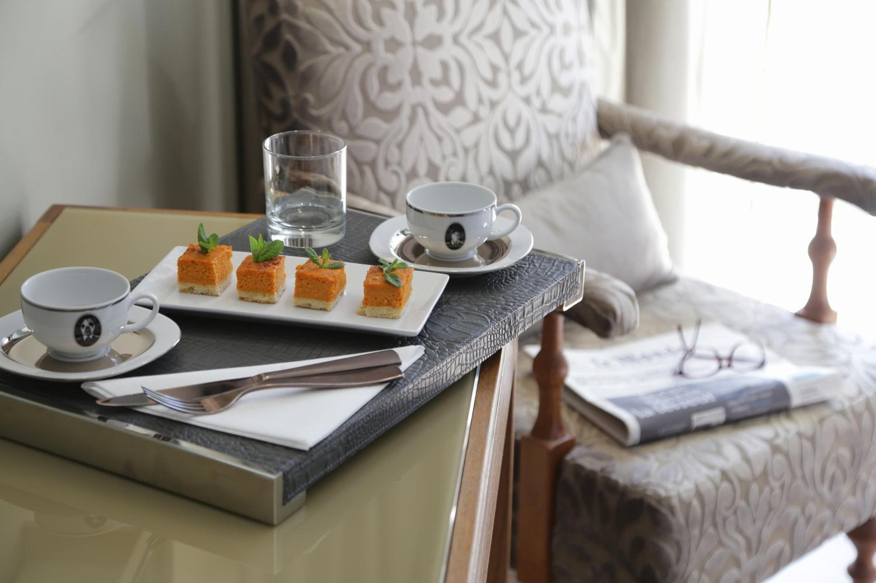 Una mesa de comedor con un plato de pasteles cuadrados, dos tazas, un vaso y cubiertos.
