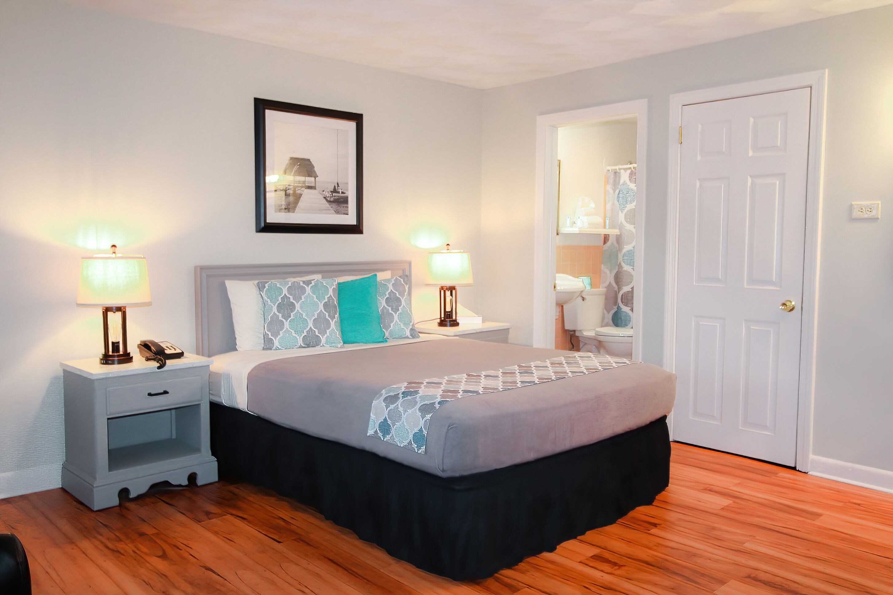 A bedroom with a bed, nightstands, a lamp, a framed picture, a door, and a bathroom visible through an open door.