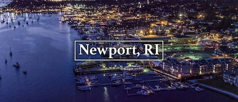 Vista aérea de Newport, Rhode Island, de noche, mostrando un puerto deportivo con numerosos barcos, edificios frente al agua y una zona urbana iluminada.