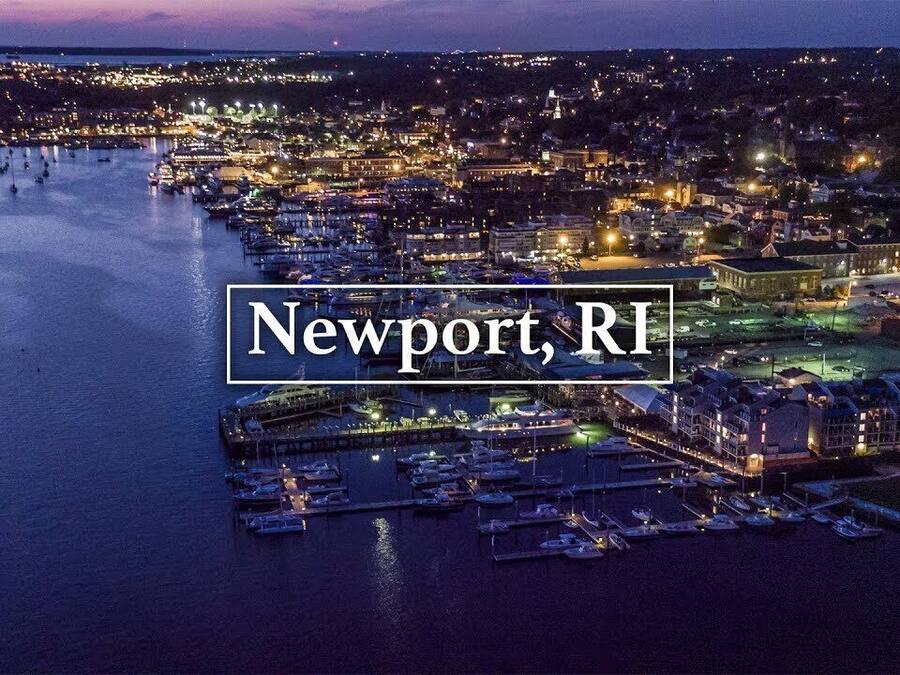 An aerial view of Newport, RI at night, featuring a marina with numerous boats, waterfront buildings, and a well-lit urban area.