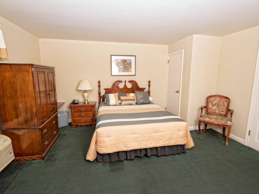 A bedroom with a large bed, a wooden wardrobe, a chair, a nightstand, and a lamp.