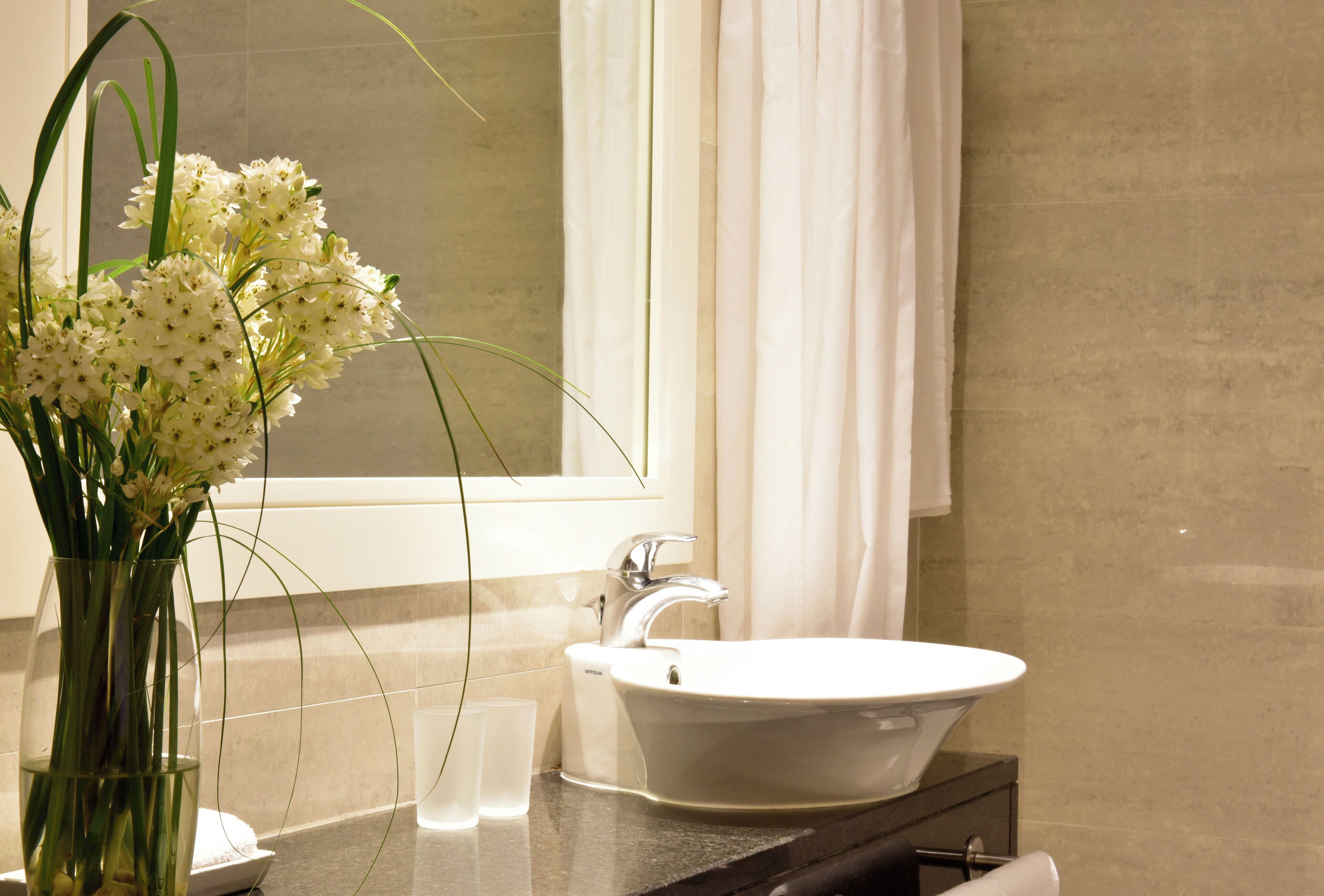 Ein Badezimmer mit einem weißen Waschbecken, einem Spiegel und einer Vase mit weißen Blumen.