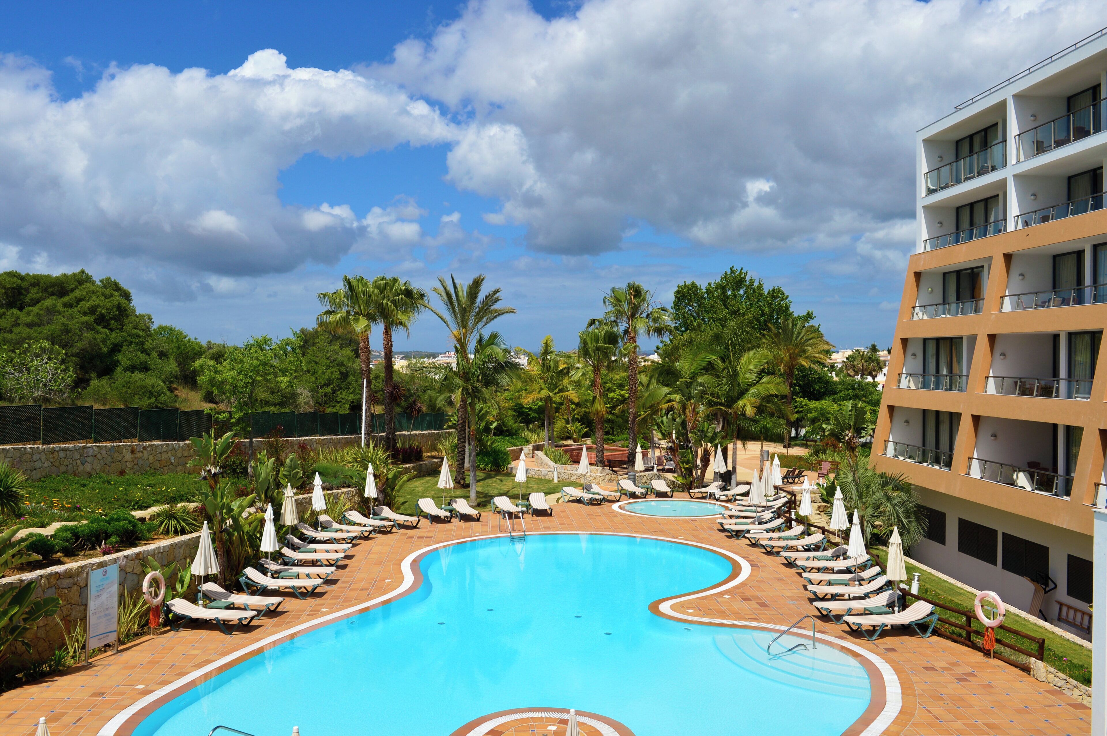A swimming pool with lounge chairs and umbrellas, surrounded by greenery and a multi-story building.
