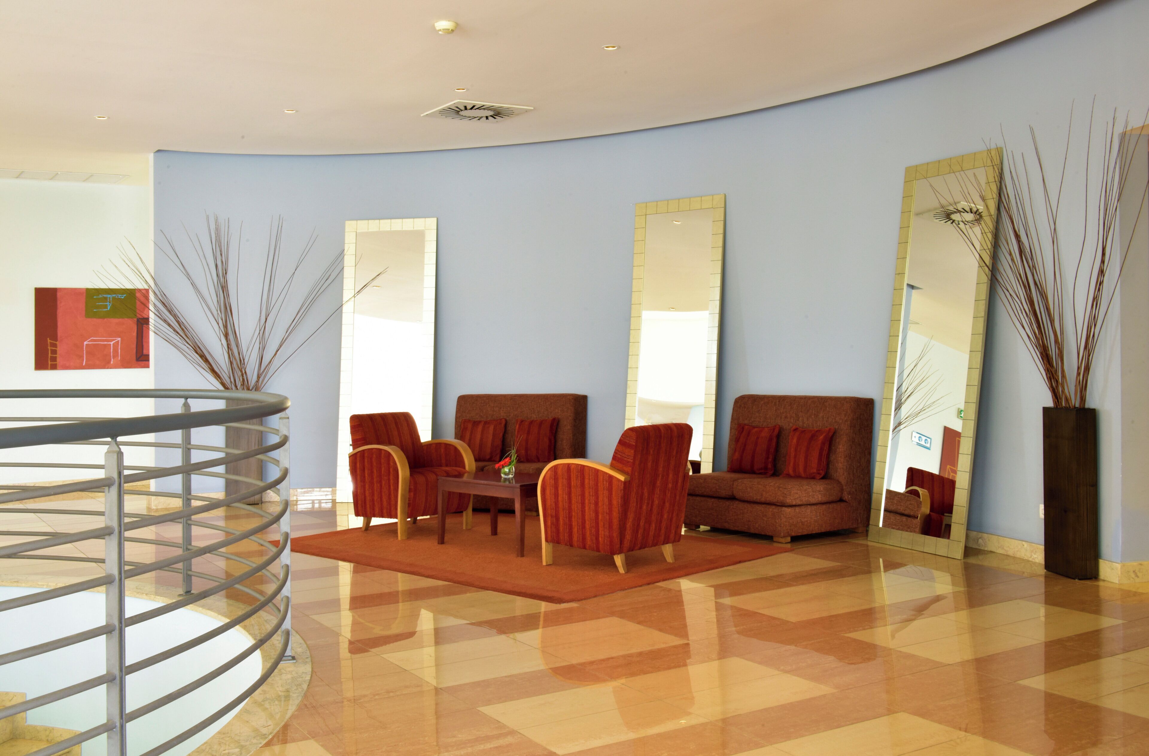 A modern lobby with a curved ceiling, a glass railing, a seating area with armchairs and a coffee table, and large mirrors on the walls.