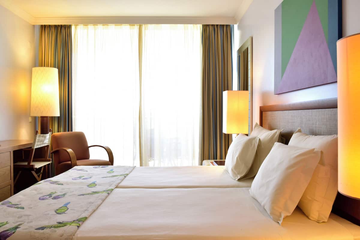 Een hotelkamer met twee bedden, een bureau, een stoel en een groot raam met gordijnen.