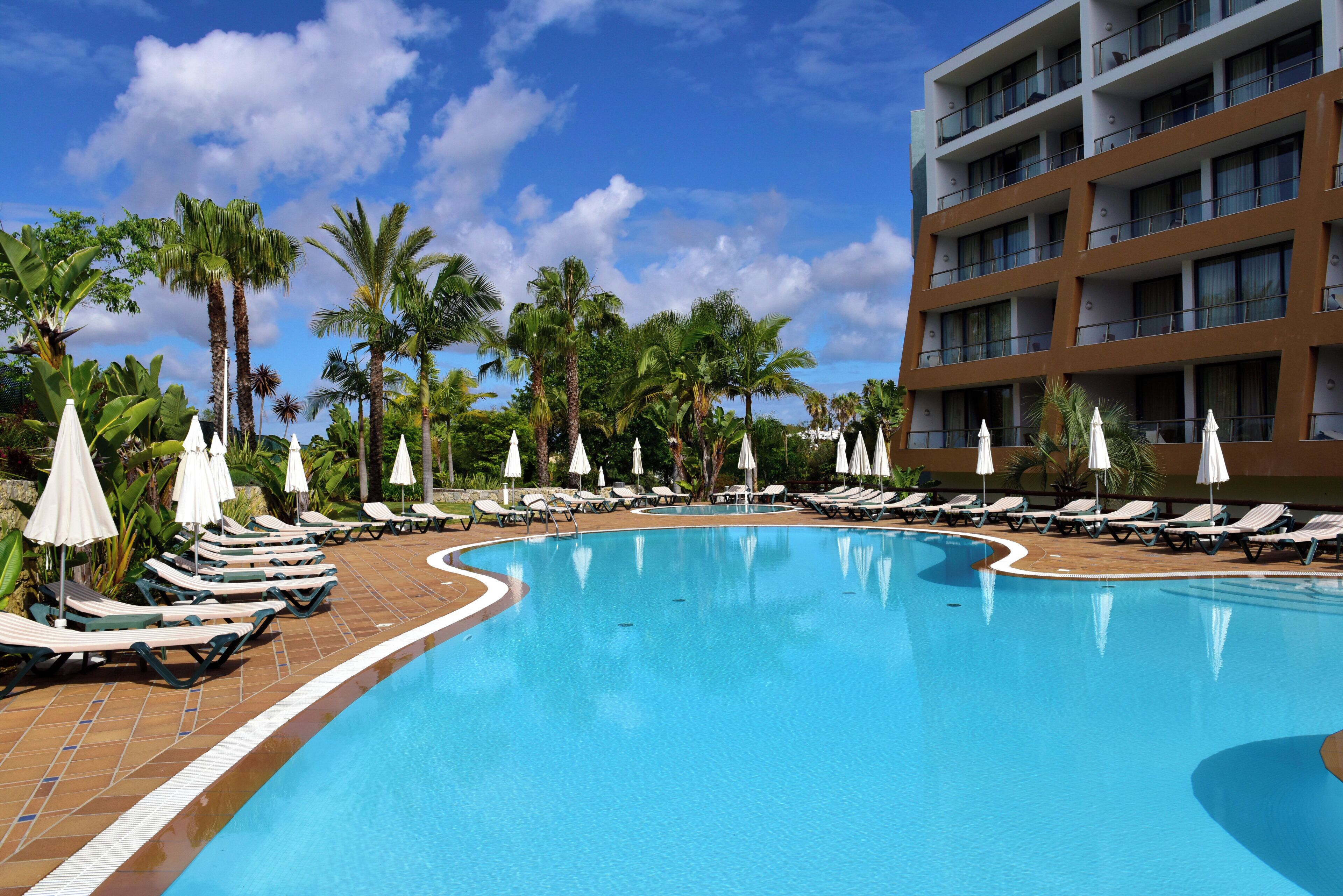 A swimming pool with lounge chairs and umbrellas, surrounded by palm trees and a multi-story building.