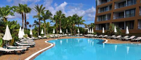 A swimming pool with lounge chairs and umbrellas, surrounded by palm trees and a multi-story building.