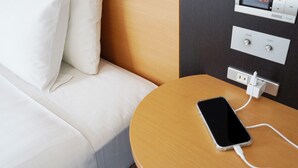Hotellihuone, jossa on sänky, yöpöytä, jossa on latauksessa oleva puhelin, digitaalinen kello ja pistorasia.