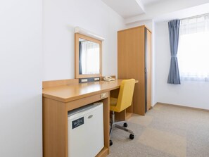 Quarto de hotel com uma secretária de madeira, uma cadeira, um eletrodoméstico branco pequeno, um espelho, um guarda-roupa e uma janela com cortinas.