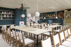 Uma sala de jantar moderna com várias mesas e cadeiras, um relógio na parede, e prateleiras com livros e objetos decorativos.