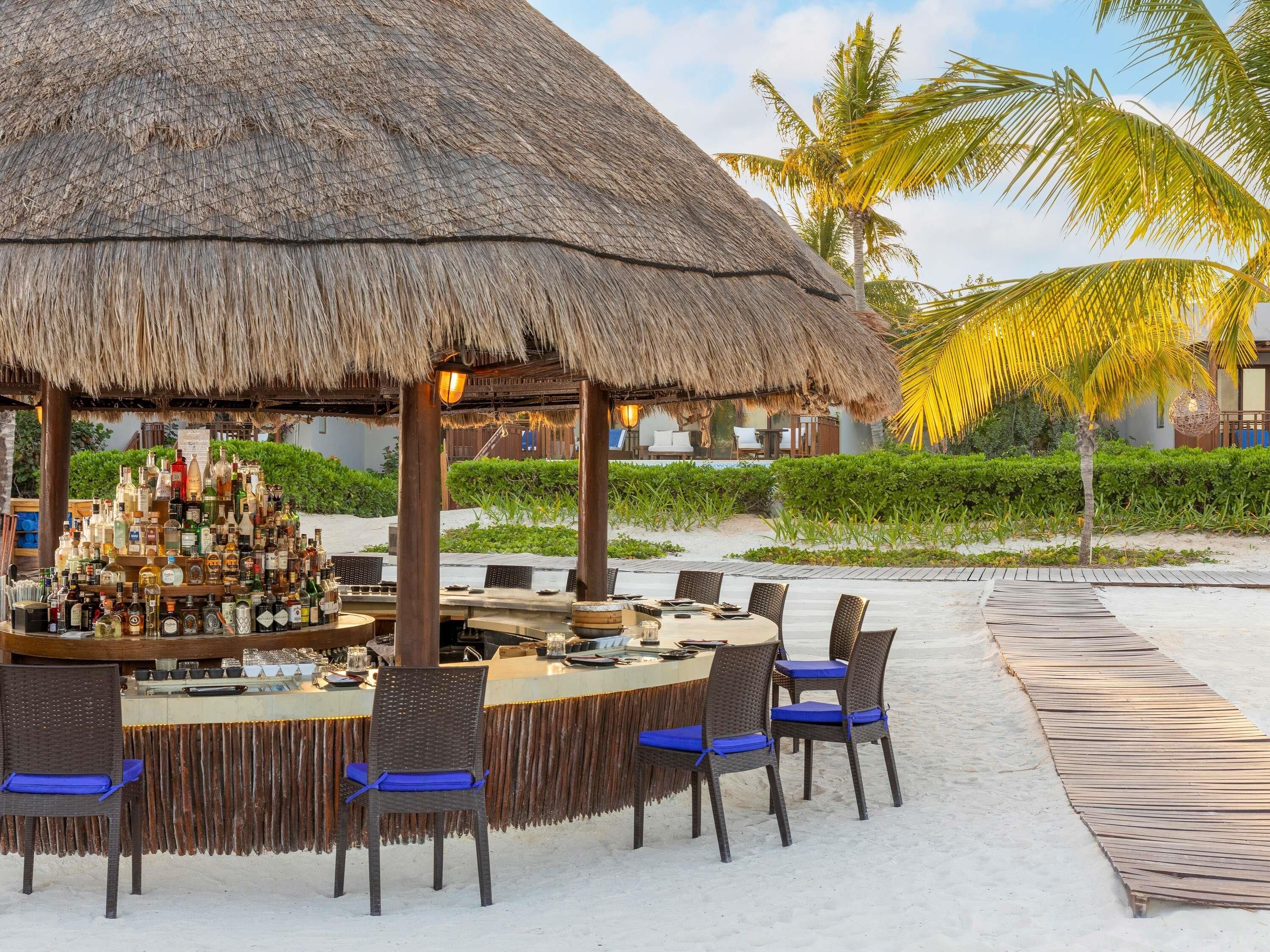 A beachside bar with a thatched roof, surrounded by wicker chairs with blue cushions, and a variety of bottles and glasses on display.