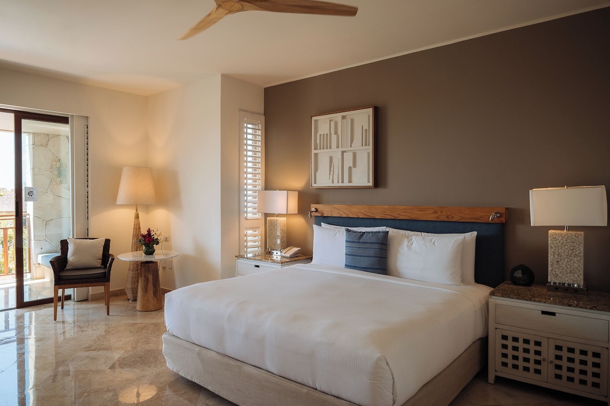 Quarto de hotel com uma cama grande, cabeceira de madeira, mesa de cabeceira com abajur, uma cadeira e vista para o exterior.