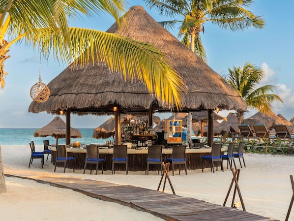 Un bar situé en bord de mer, doté de parasols en chaume, de chaises longues et d’une terrasse en bois qui mène à la plage.