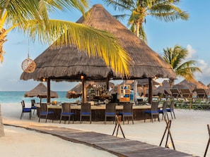 A beachside bar with thatched umbrellas, lounge chairs, and a wooden deck leading to the sand.