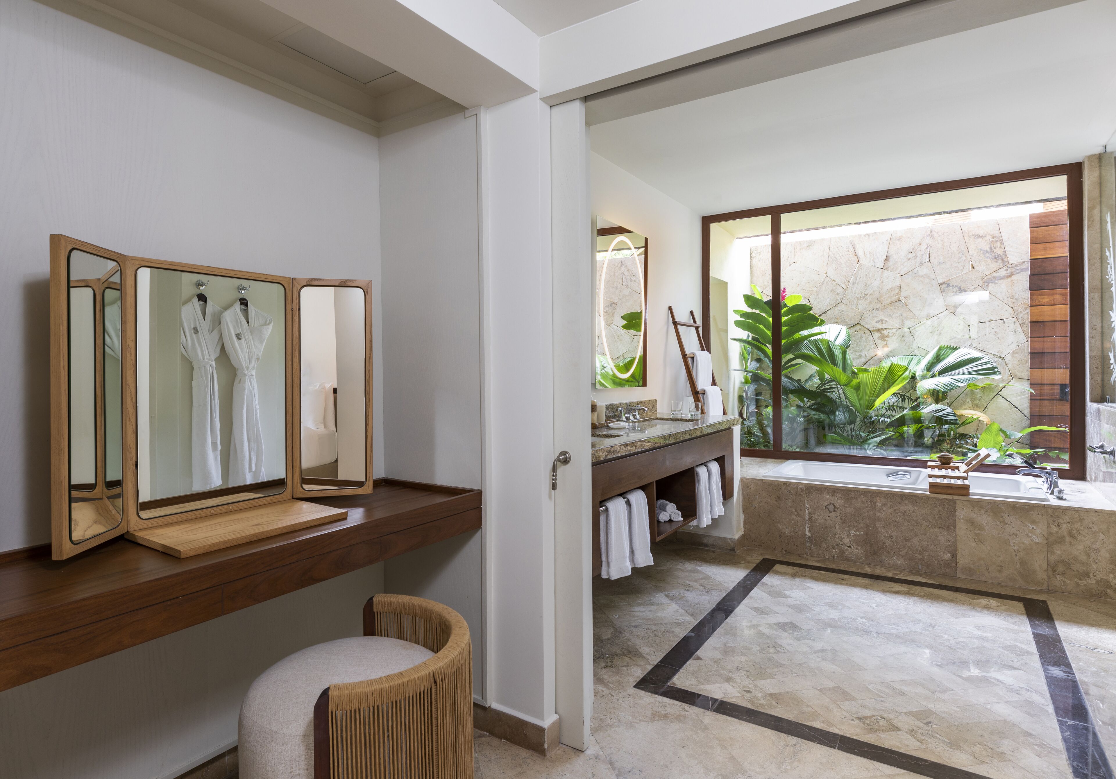 A modern bathroom with a wooden vanity, a large mirror, a bathtub, and a view of a stone wall and plants.