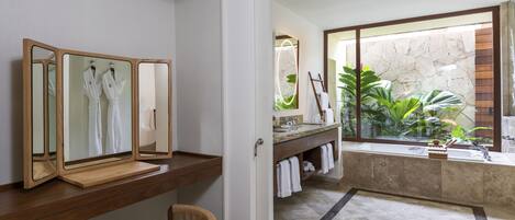 A modern bathroom with a wooden vanity, a large mirror, a bathtub, and a view of a stone wall and plants.