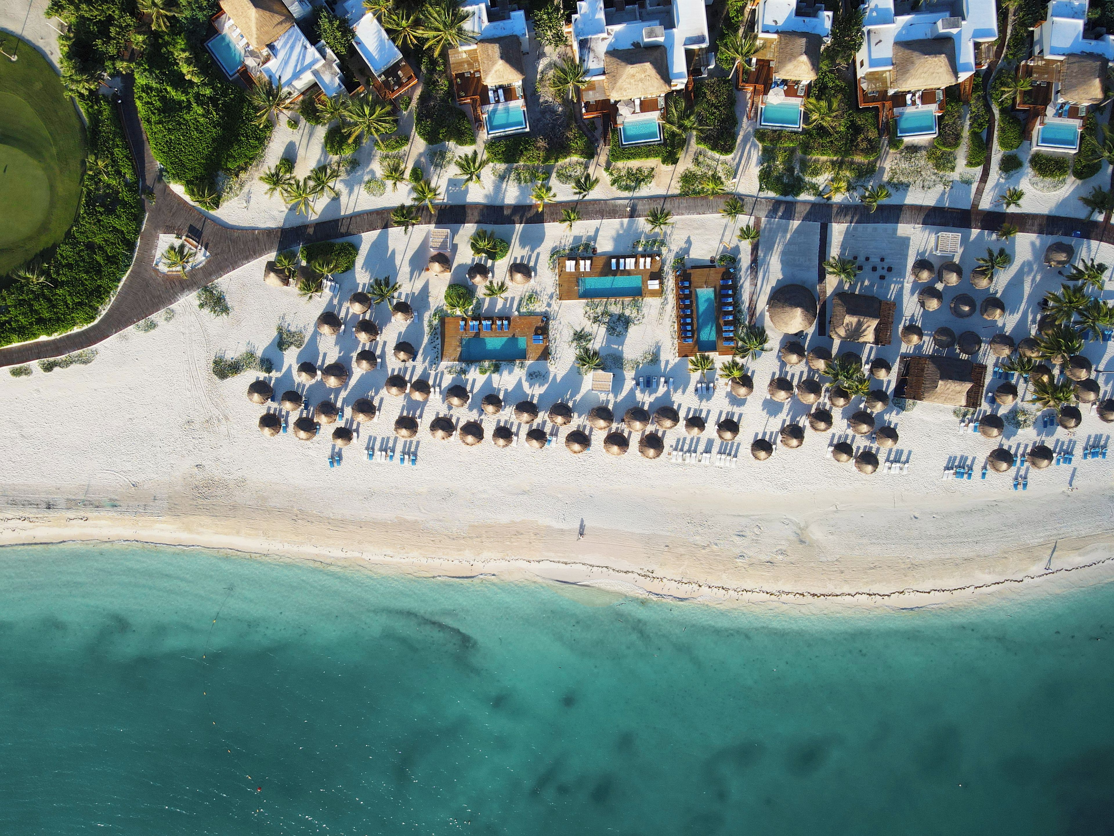Vista aérea de un complejo turístico en la playa con múltiples piscinas, tumbonas y una playa de arena.