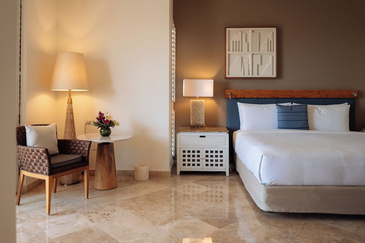 A hotel room with a bed, a chair, a small table with a lamp, a nightstand, and a wall art piece.