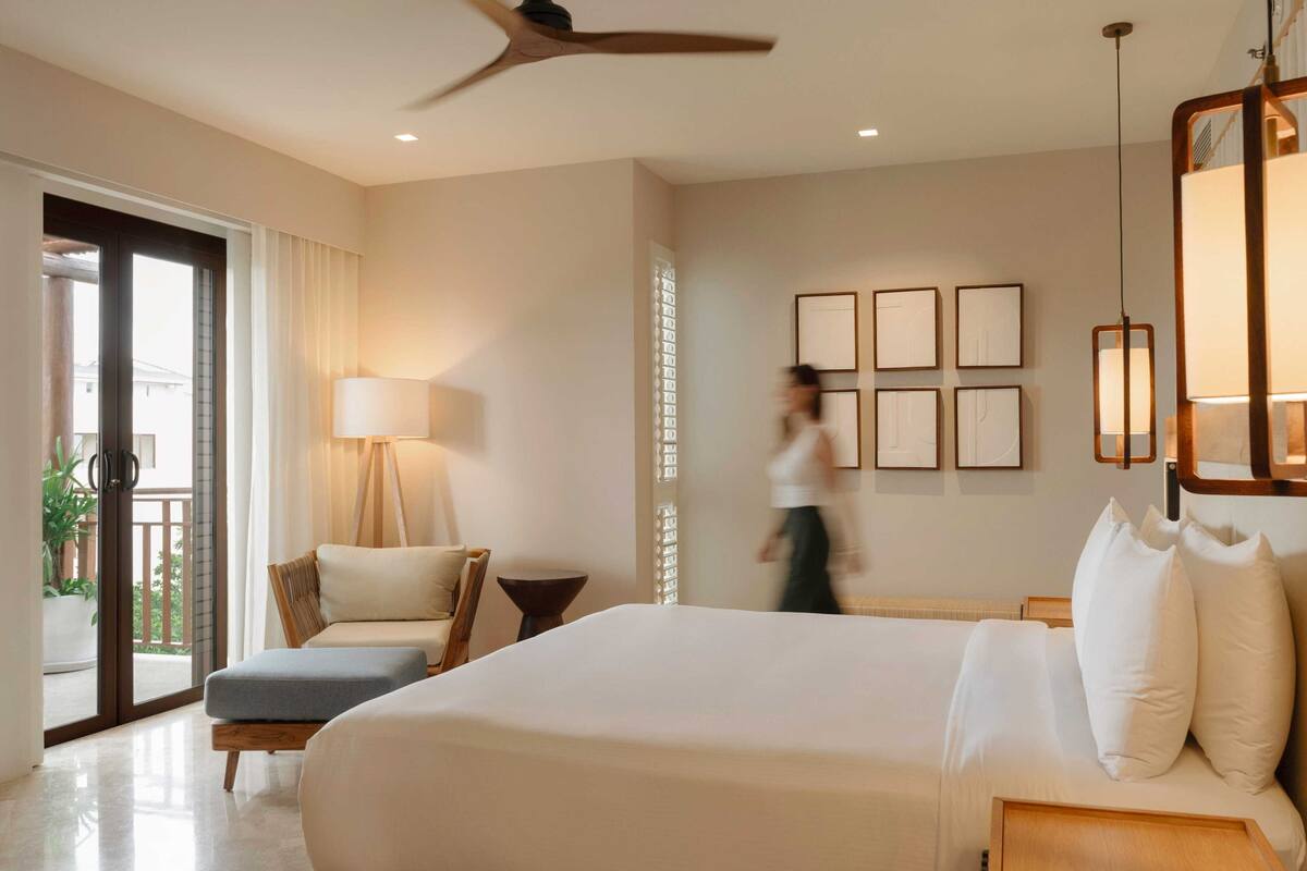A hotel room with a bed, a chair, a table, a lamp, and a balcony.