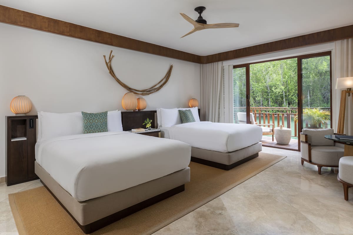 A modern hotel room with two beds, a ceiling fan, a large window with a view of greenery, and a balcony with a wooden railing.