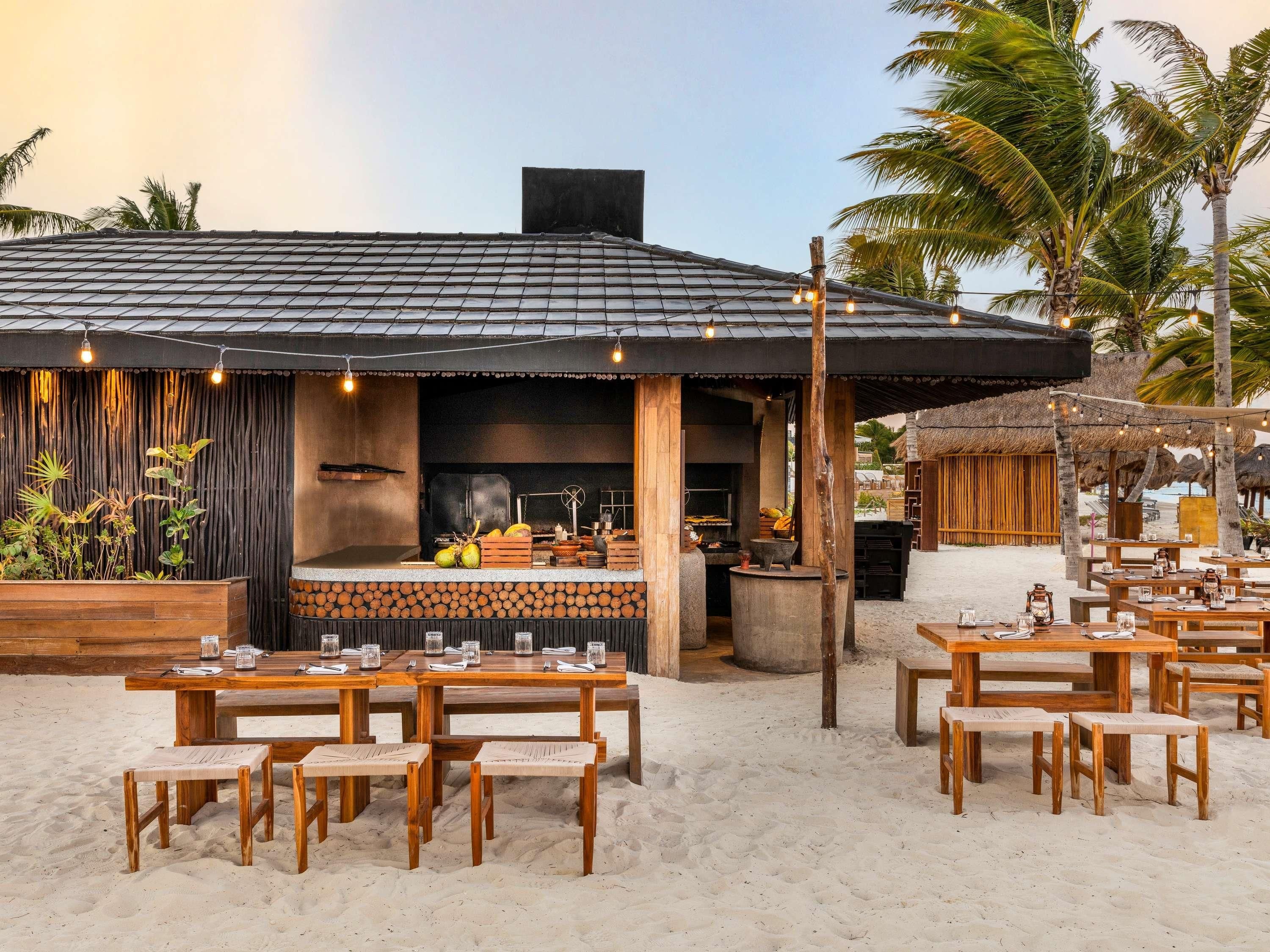 A beachside restaurant with outdoor seating, a grill, and a thatched-roof structure.